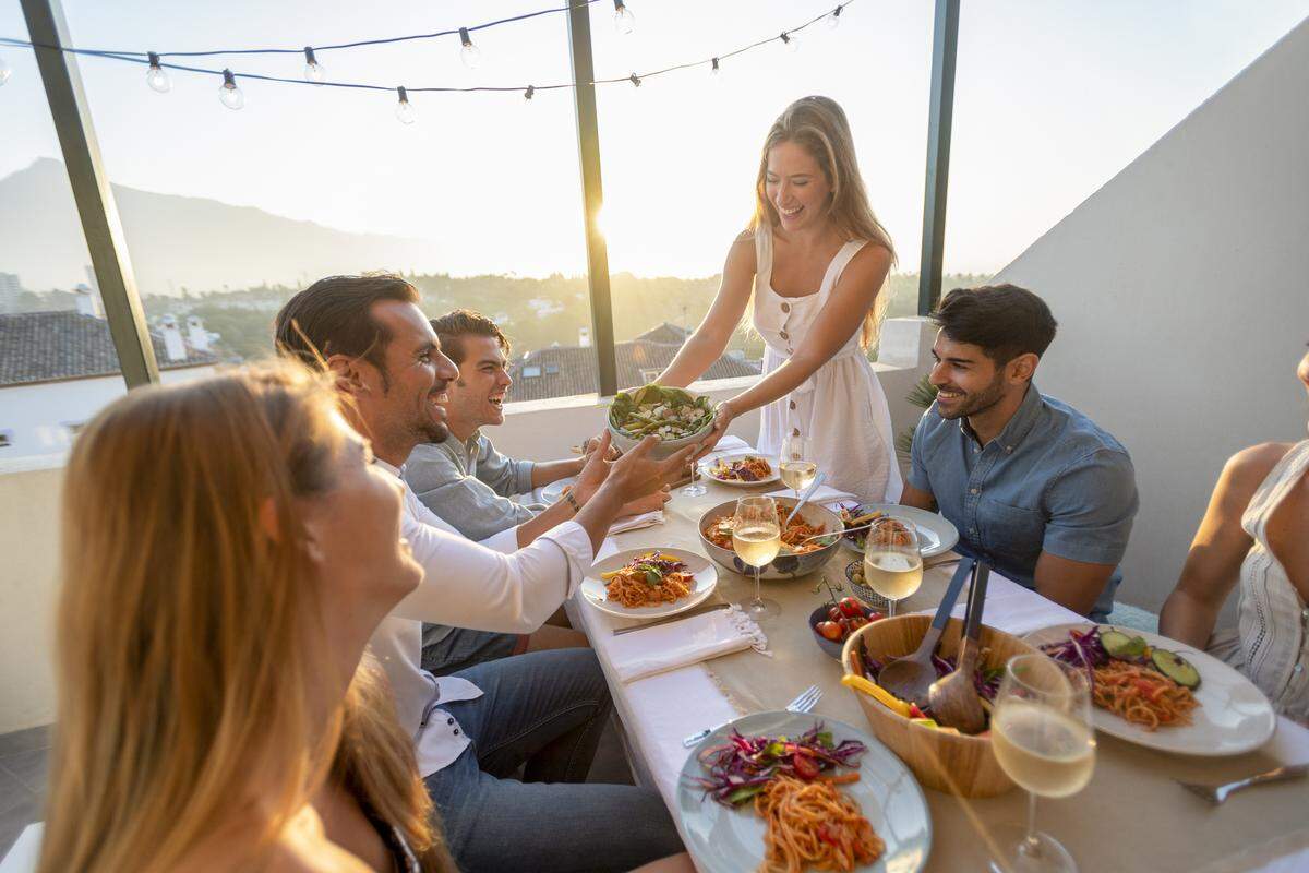 Friends and family passing salad on a table at sunset. There is spaghetti Bolognese on some of the plates on the table. There are glasses of wine and other food on the table; everyone is having fun laughing and smiling