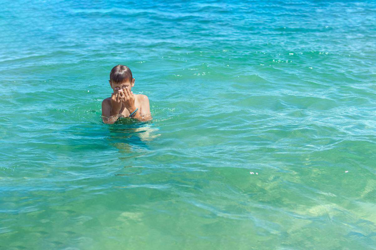 A young boy plays in the ocean covering his face.