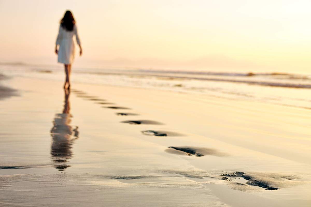 Full length of woman walking on wet sea shore at sunset. Female in sundress is leaving her footprints on sand. 