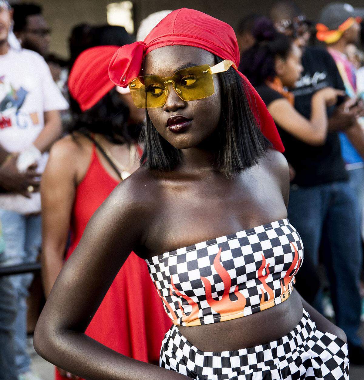 A medium-close shot features a Black person with a deep complexion posing confidently in a crowded outdoor setting, styled in a bold, streetwear-inspired outfit and durag, looking slightly over one shoulder toward the right of the frame.