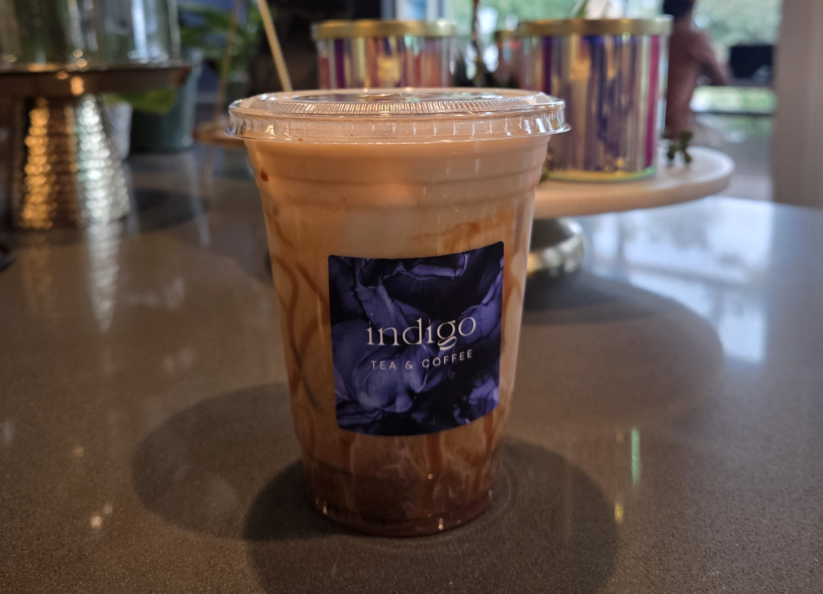 A close-up shot features a clear plastic cup of iced, caramel-swirled coffee or tea with an “indigo tea & coffee” label. The beverage is sitting on a gray counter, with a tiered stand holding colorful, reflective candle holders blurred in the background.