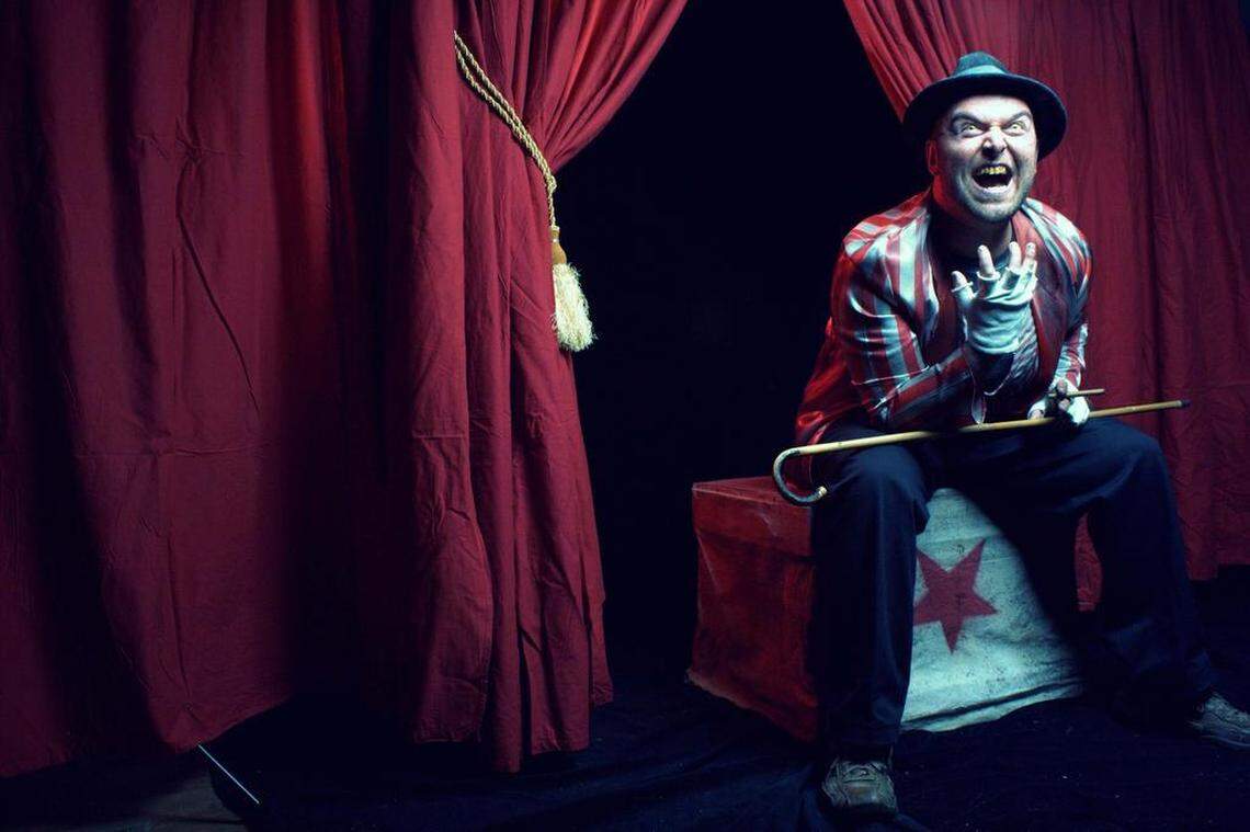 A menacing-looking man dressed in a striped, reddish-white circus ringmaster or clown jacket, dark pants, a fedora, and white gloves sits on a painted box with a red star. He is holding a walking stick or cane, leaning forward, and has a wide, frightening, teeth-baring grin. He is framed by heavy, red velvet curtains on a dark background.