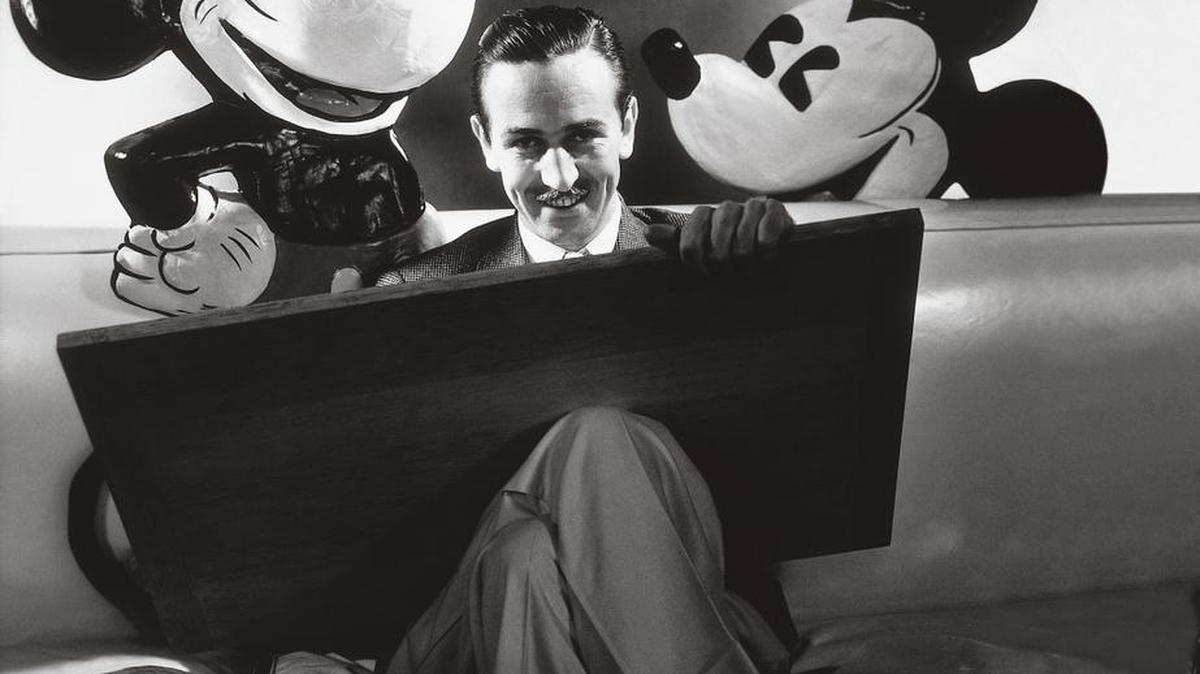 
Walt Disney seated with drawing board on his lap and representations of his creations Mickey and Minnie Mouse, ca. October 1933. 
