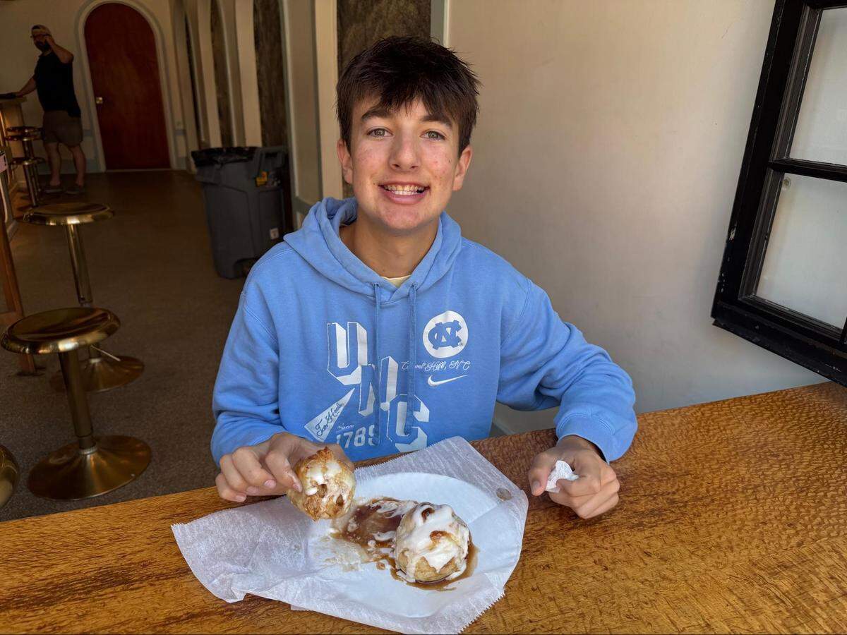 An eye-level, medium close-up shot of a smiling teenager sitting at a wooden counter, wearing a light blue UNC Chapel Hill hooded sweatshirt and holding a small pastry roll topped with white icing in his right hand. On a white paper plate lined with wax paper in front sits another pastry roll covered in thick white icing and a sweet, brown glaze. The blurred background shows a restaurant hallway with gold barstools and a person standing in the distance.