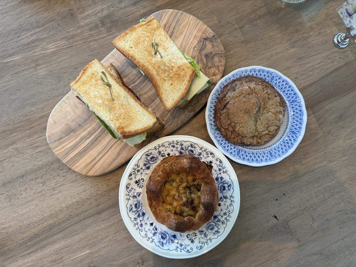 A top-down view of a selection of bakery items arranged on a smooth, grey-brown wooden table. On the top left, two halves of a toasted sandwich with visible cheese and greens are served on an oval olive-wood serving board. To the right is a large, golden-brown crinkle cookie with a sugary dusting on a blue and white patterned plate. At the bottom center, a round, flaky savory pastry filled with a golden, baked cheese topping rests on a white plate adorned with an intricate blue floral design.