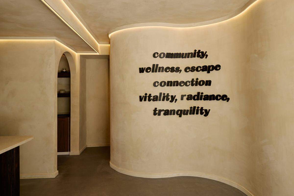 A minimalist interior space with light-colored, curved walls and soft, recessed lighting. Black, three-dimensional letters are mounted on the main wall, spelling out a list of words: “community,” “wellness, escape,” “connection,” and “vitality, radiance, tranquility.” A rounded archway in the background leads to another room with wooden shelving.