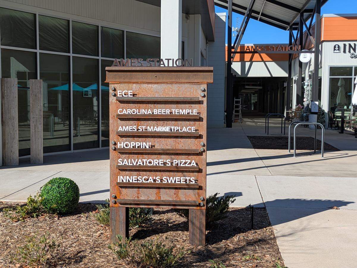 A weathered, rusted metal directory sign for “Ames Station” standing outdoors. The sign lists local businesses including ECE, Carolina Beer Temple, Ames St Marketplace, Hoppin’, Salvatore’s Pizza, and Innesca’s Sweets.