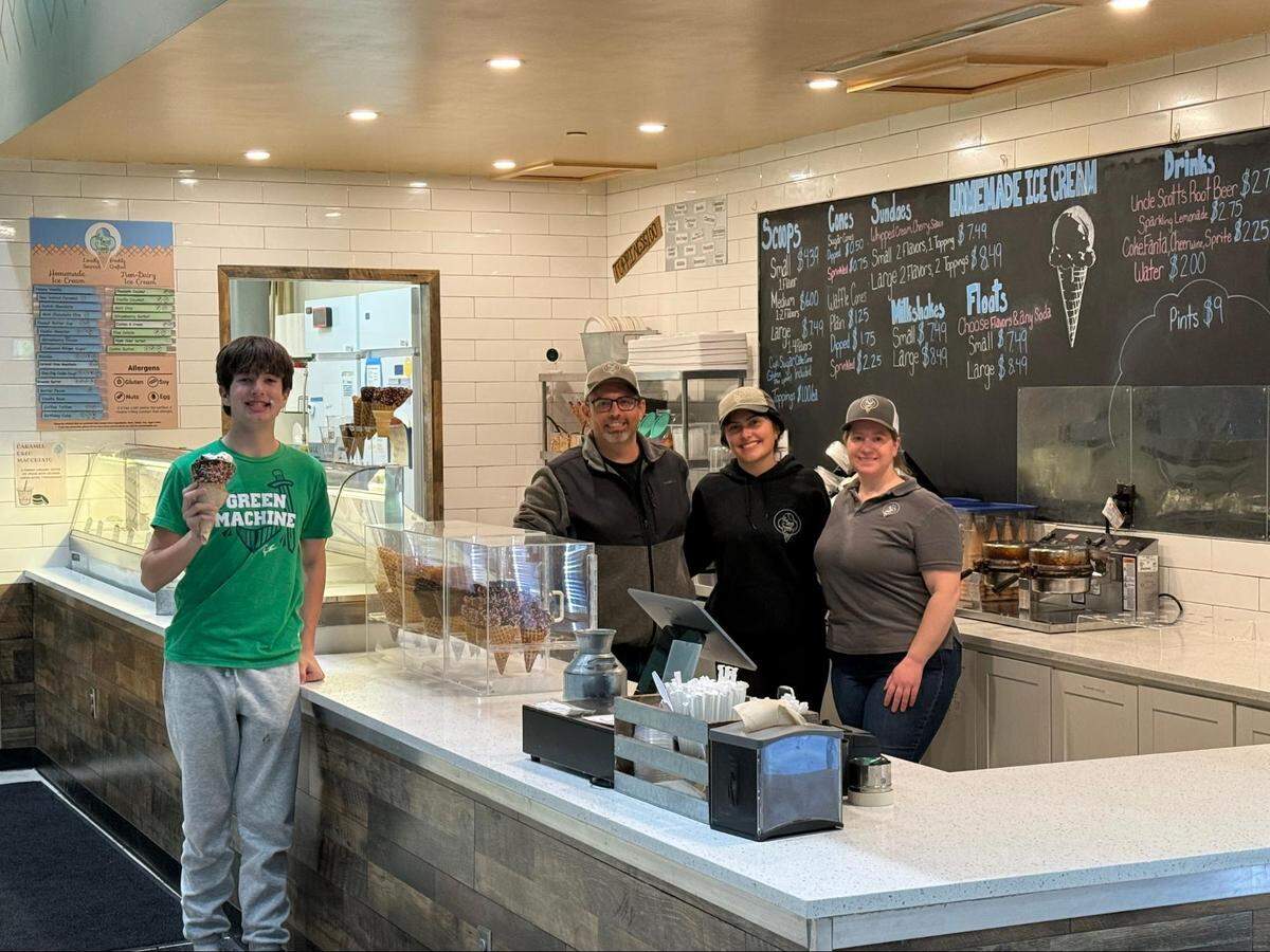 Inside an ice cream shop, a young person stands in front of the counter holding a chocolate-dipped waffle cone covered in sprinkles. They are wearing a green t-shirt that says “GREEN MACHINE” and grey sweatpants. Behind the white counter, three employees stand together smiling at the camera. The shop features white subway tile walls, a glass ice cream display case, and a large black chalkboard menu displaying prices and flavors.