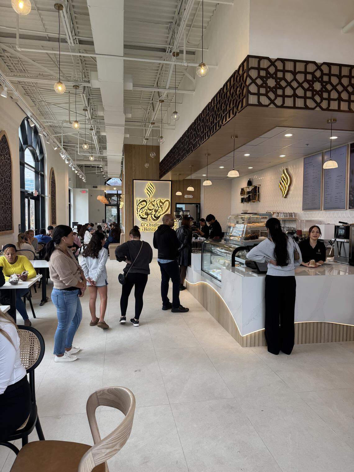 Interior view of a brightly lit café with modern white counters and high ceilings. Customers are standing in line near the service counter, and others are seated at tables near arched windows.