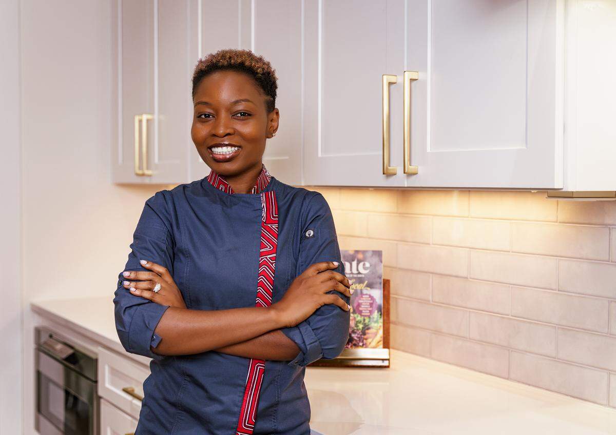 A smiling chef wearing a dark blue chef’s coat with a red and white patterned trim along the collar and zipper stands with arms crossed in a bright, modern kitchen. The chef has short, reddish-brown hair and is wearing a ring on the left hand. The background features white shaker-style cabinets with gold handles, under-cabinet lighting, a light-colored subway tile backsplash, and a white countertop. A cookbook is visible on the counter.
