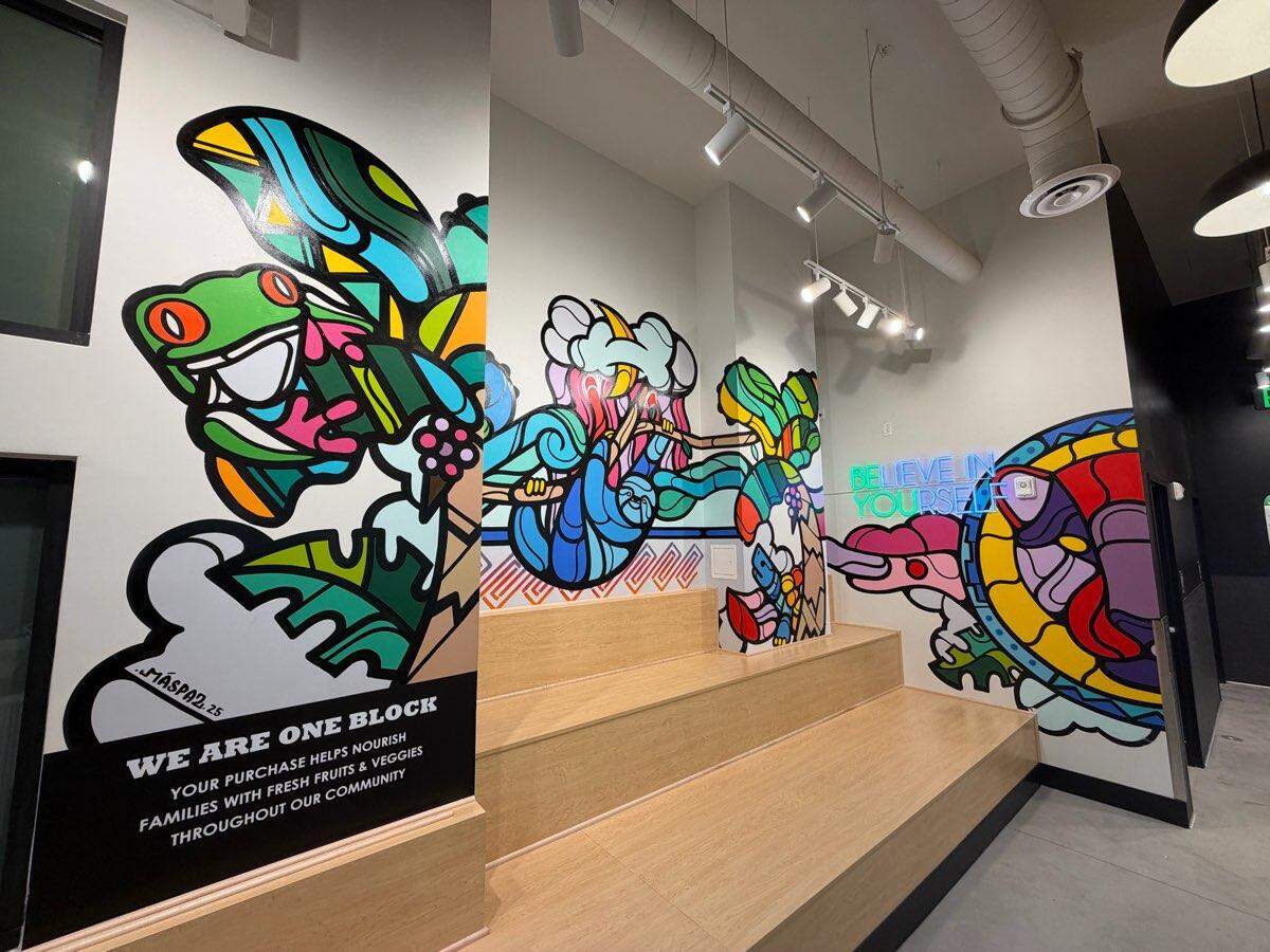 An interior view of South Block Ballantyne featuring a vibrant, multi-colored mural with stylized animals including a frog, a sloth, and a turtle. The space includes tiered light-wood bleacher seating and a neon sign on the wall that reads “BELIEVE IN YOURSELF” in bright blue and green.