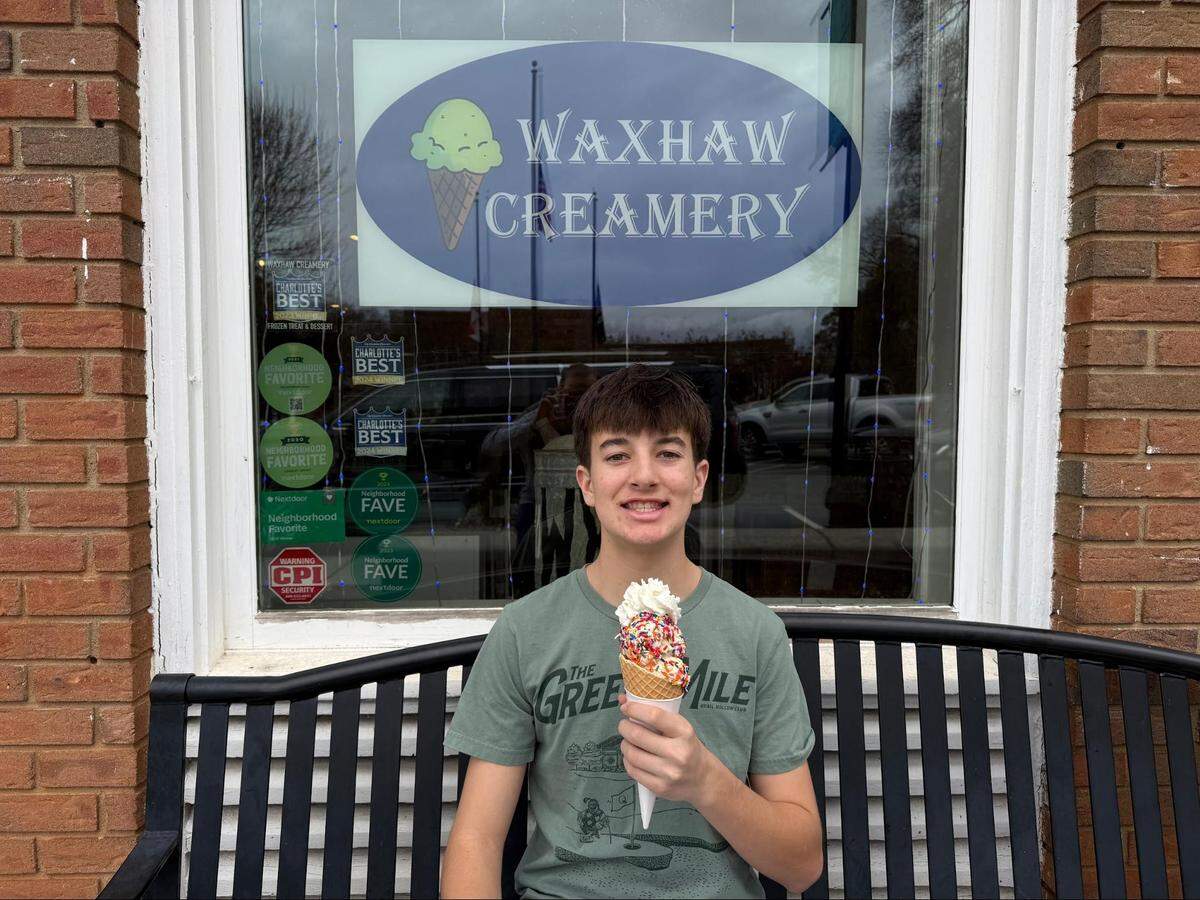 A teenager sits on a black metal bench in front of the Waxhaw Creamery window, holding a large waffle cone topped with colorful sprinkles and whipped cream. The window displays several “Charlotte’s Best” and “Neighborhood Favorite” award stickers.