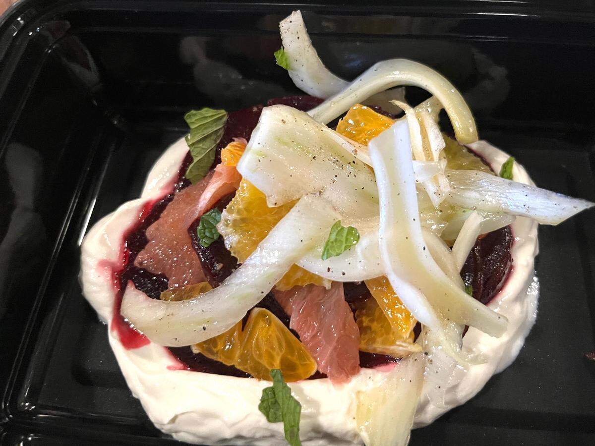 A close-up, overhead view of a colorful salad served in a black plastic takeaway container. The dish features a layer of creamy white base (likely yogurt, cream, or cheese) topped with dark red beet puree, segments of bright orange and pink grapefruit, and thin, curving slices of white fennel bulb. The dish is garnished with small mint leaves and a light dusting of black pepper.