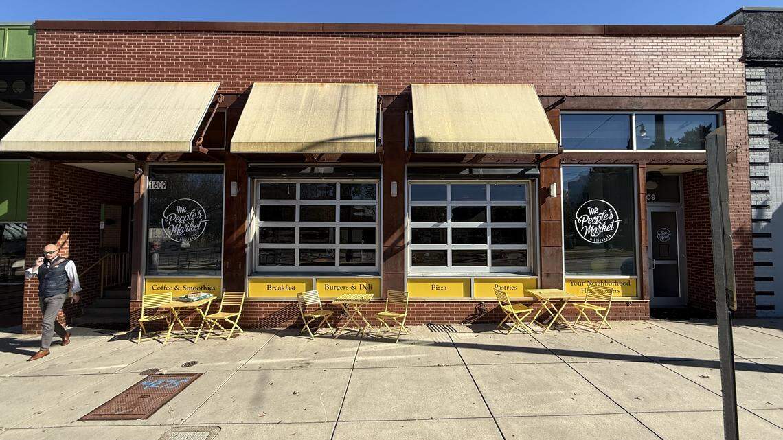 A straight-on, eye-level wide shot capturing the full facade of “The People’s Market” from across the street. The one-story, red brick building has three large, weathered tan awnings above two central, glass-paneled garage doors and side windows. Several yellow metal tables and folding chairs are set up on the concrete sidewalk for outdoor seating. On the far left, a man is walking past the building on a bright, sunny day.