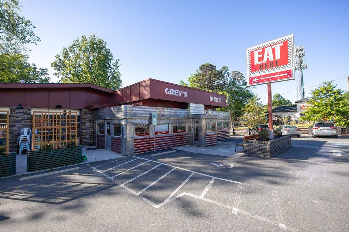 An exterior shot of Grey’s Diner, a classic mid-century style diner with a stainless steel and red ribbed facade. A large vintage-style sign stands tall in the parking lot with a red background and white text that reads “EAT HERE” above a flashing arrow. The building sits under a clear blue sky, surrounded by green trees and a paved parking lot with a handicap space in the foreground.