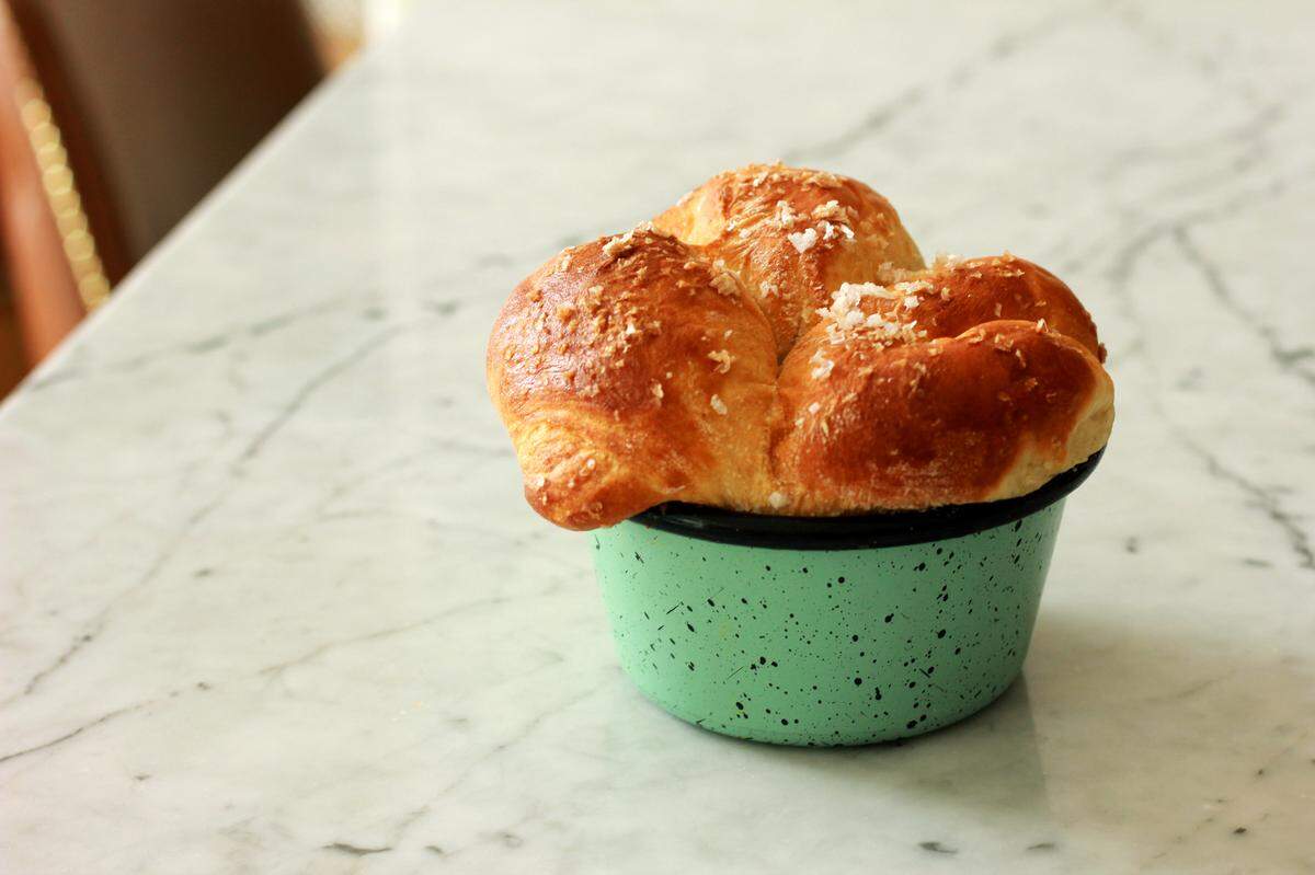 A close-up shot of a golden-brown, freshly baked milk bread, possibly a pull-apart loaf or rolls, rising out of a speckled mint green enamel mug. Large crystals of salt are visible sprinkled over the top of the bread, which has a shiny, golden crust. The mug sits on a white marble countertop, with a blurred background suggesting an indoor setting.
