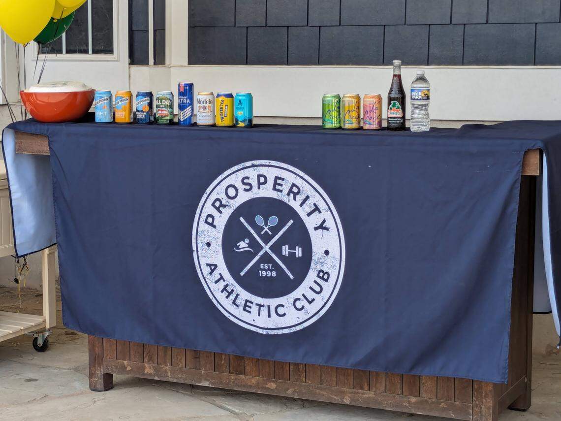 A wooden table is set up as a bar for a gathering. It’s covered by a dark blue cloth with a large, white, circular logo for the “Prosperity Athletic Club.” A variety of canned beers, seltzers, and bottled water are lined up on the tabletop next to an orange punch bowl.
