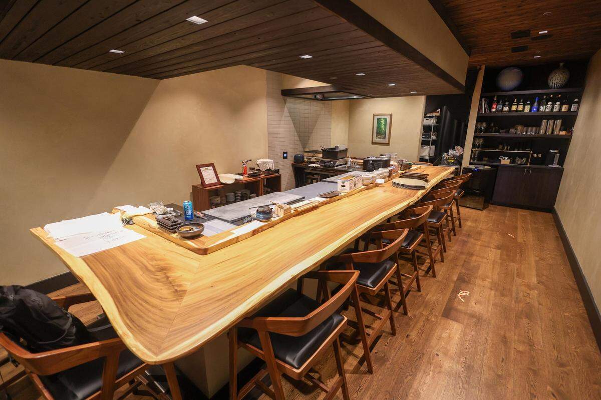 The interior of an upscale sushi restaurant, featuring a long, L-shaped, live-edge wooden sushi bar lined with modern dark wood chairs. The room has a minimalist aesthetic with wood-paneled ceilings.