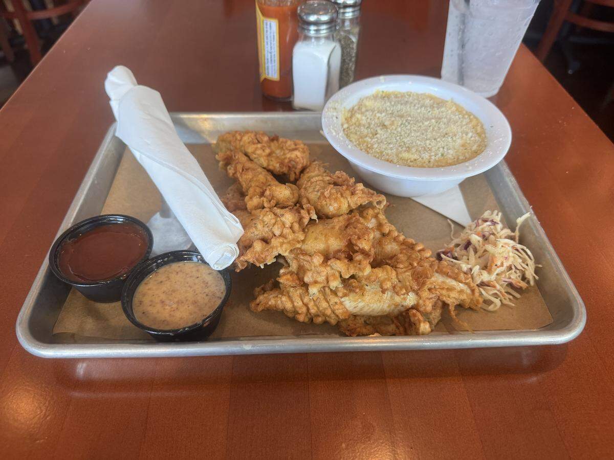 A top-down view of a serving of crispy fried chicken tenders on a metal tray lined with brown paper. The tray also holds two dipping sauces in black plastic cups, a small portion of coleslaw, and a large bowl of mac and cheese with a breadcrumb topping. A rolled-up napkin and silverware are on the tray, and salt and pepper shakers and a hot sauce bottle are in the background.