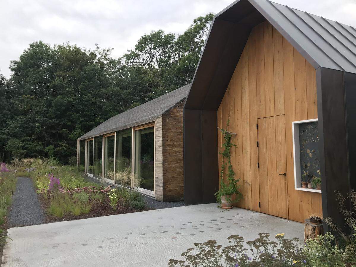 An exterior view of Noma 2.0 in Copenhagen, featuring a modern, minimalist wood-paneled building with a steep gable and dark metal trim. A gravel path winds through a wild, naturalistic garden with wildflowers leading toward a long, glass-walled structure with stacked-wood siding under a cloudy sky.