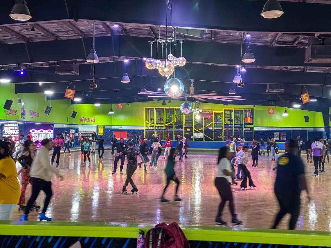 A wide, bustling shot of a crowded indoor roller skating rink. Dozens of people are skating on the polished, reflective wooden floor under several large disco balls hanging from the ceiling. The background walls are painted a vibrant lime green, with signs for a “Game Room” and “Rental” counter visible. The photo captures the active, social atmosphere of the rink, with many skaters blurred in motion.