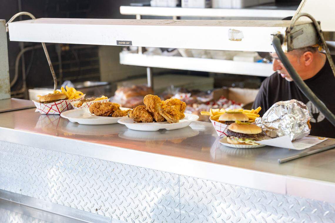 A close-up, eye-level shot of a stainless steel restaurant counter lined with food ready for service. Several paper plates and baskets hold golden-brown fried chicken, burgers, and french fries under a warming lamp. A restaurant worker is partially visible in the background on the right.