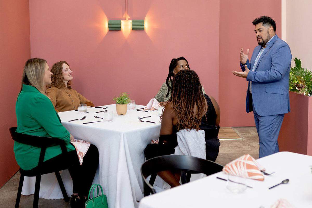 A chef in a blue suit stands and speaks to four seated diners at a white-clothed table. The setting features a modern coral-pink background with a minimalist green wall sconce. The diners, including women in a green blazer and a brown button-down, look on as the chef gestures with his hands during the presentation.