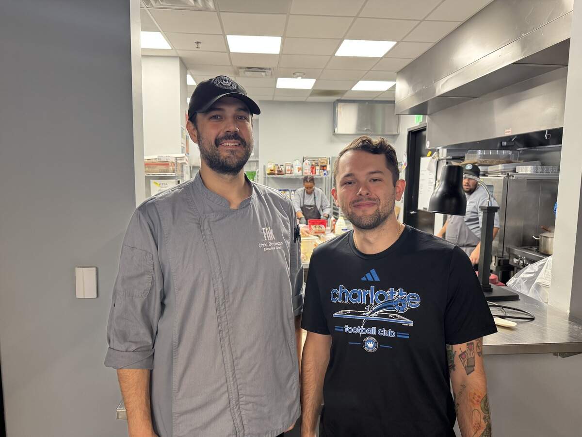 Two men smiling side-by-side inside the doorway of a brightly lit commercial kitchen. The man on the left is wearing a grey chef’s jacket with “Flik Executive Chef” embroidered on the chest and a black baseball cap. The man on the right is wearing a black Charlotte Football Club T-shirt. Stainless steel appliances, shelves, and another kitchen worker are visible in the background.