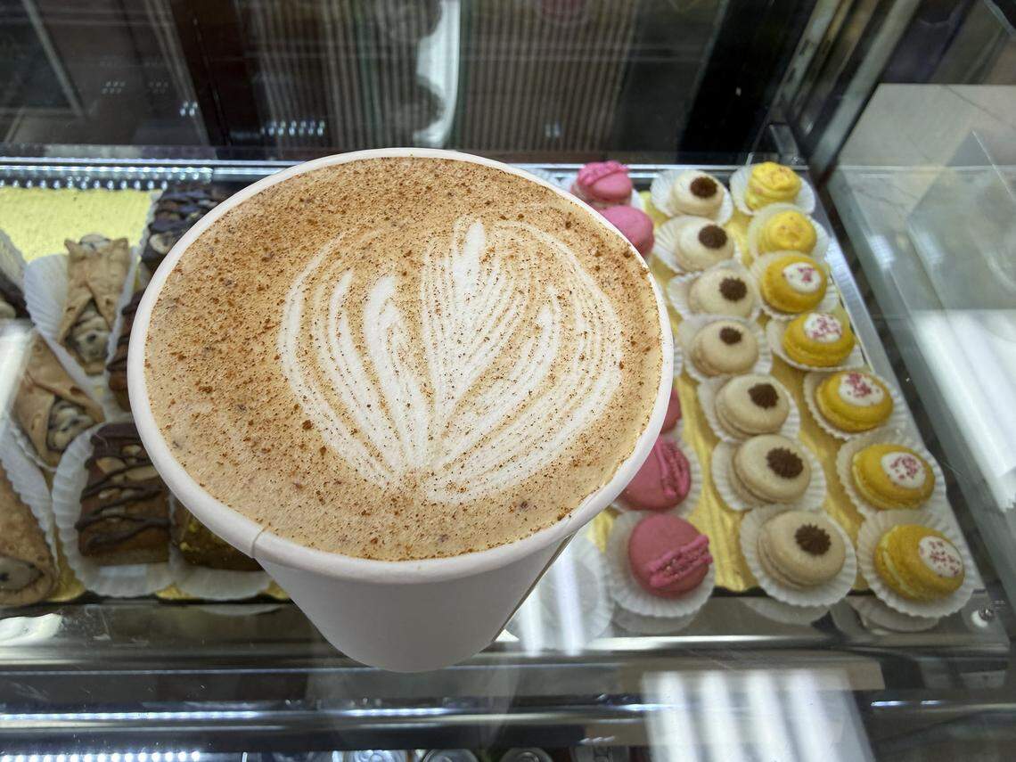 A high-angle, close-up shot of a latte in a white paper cup, featuring detailed foam art in a leaf pattern and a dusting of cinnamon. In the blurred background, rows of colorful macarons and cannolis are visible inside a bakery display case.