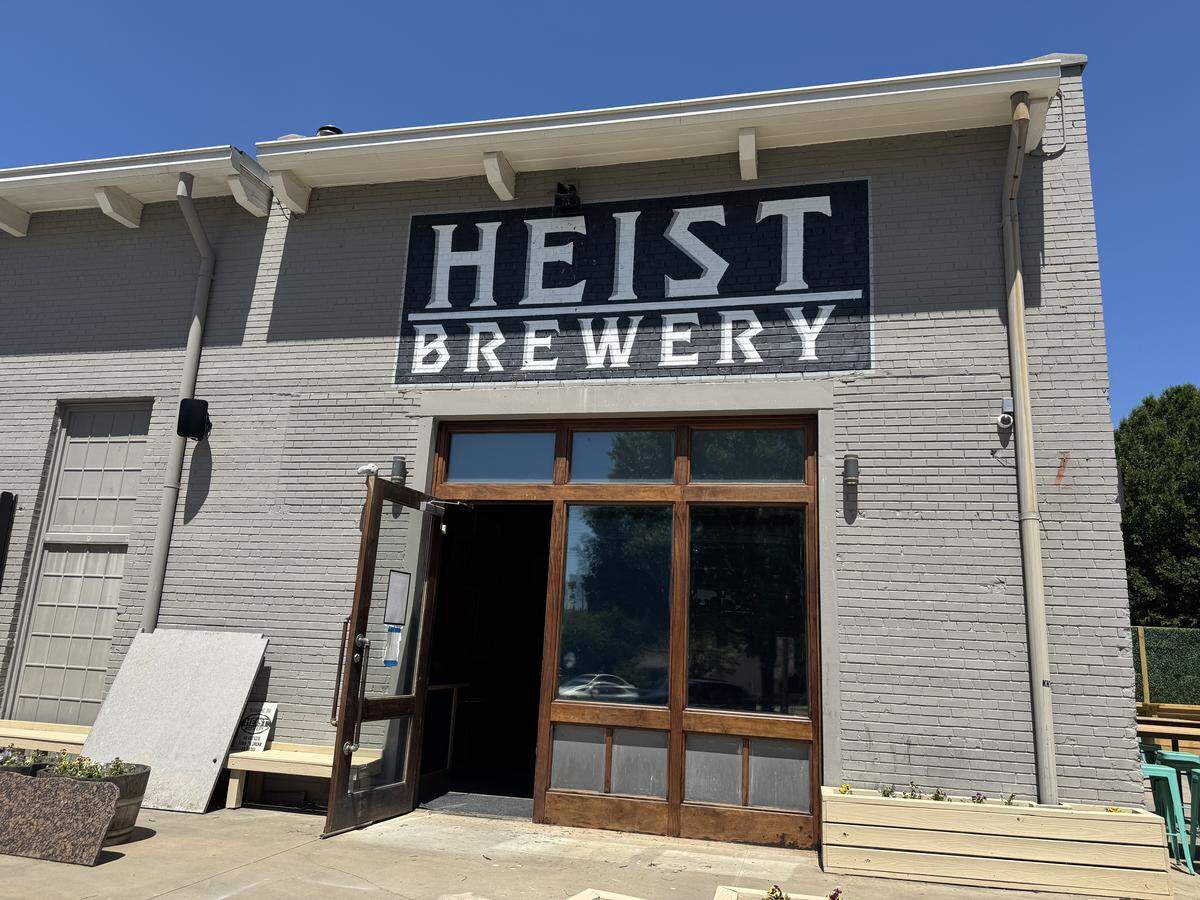 A wide, eye-level shot of the exterior entrance to Heist Brewery. The building is made of light gray painted brick. Above the large wooden-framed glass doors, a prominent black sign with white lettering reads “HEIST BREWERY.” To the left of the door, a few stone slabs lean against the wall next to a wooden bench. The sky above is a clear, bright blue.