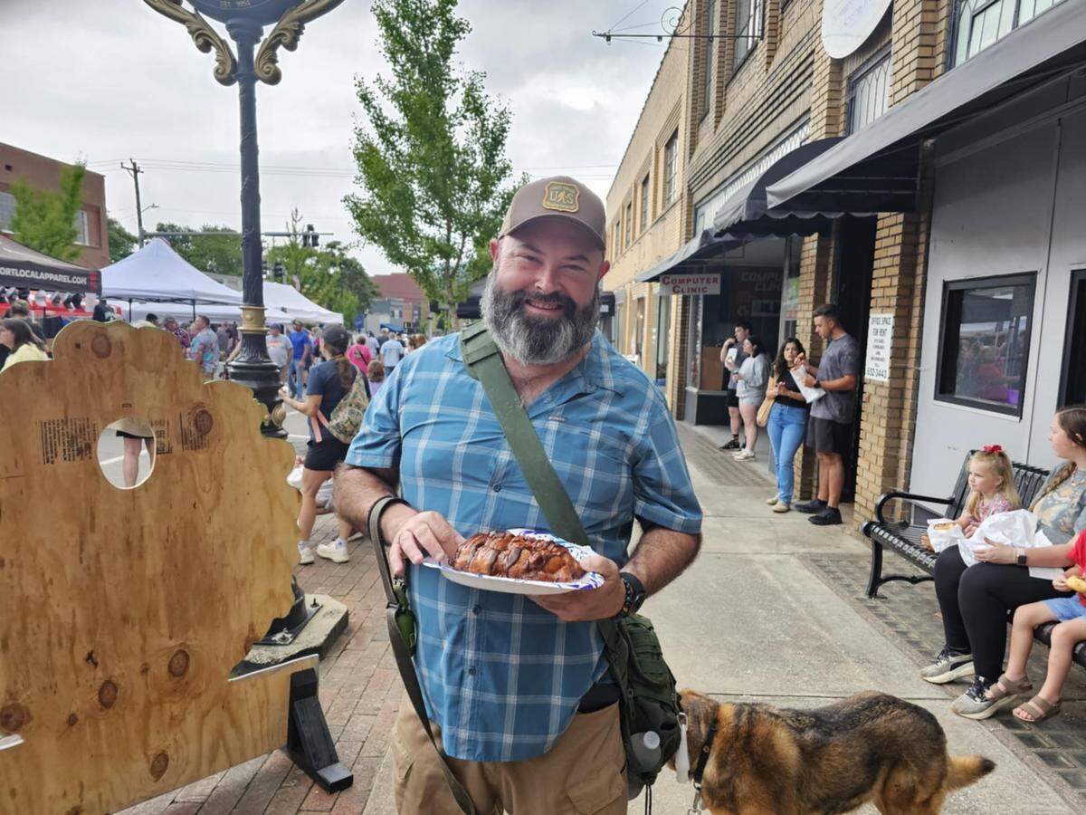 A man in a plaid shirt and cap smiles for a photo in the middle of a street fair. He holds a paper plate with a large, golden-brown pastry called a “Bigfoot Cuban Bear.” A dog on a leash is next to him, and in the background are brick buildings, vendor tents, and other attendees.