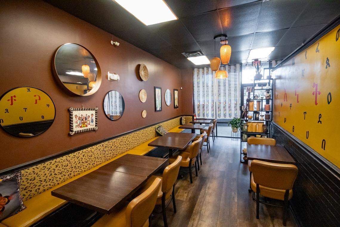 A perspective shot looking down the length of the dining area. The space is warm and inviting, featuring chocolate-brown walls decorated with circular mirrors and traditional woven plates. A long yellow banquette with leopard-print backing runs along the left wall, paired with dark wood tables and tan leather chairs. The lighting is soft, provided by woven basket pendant lights and natural light from the front window.