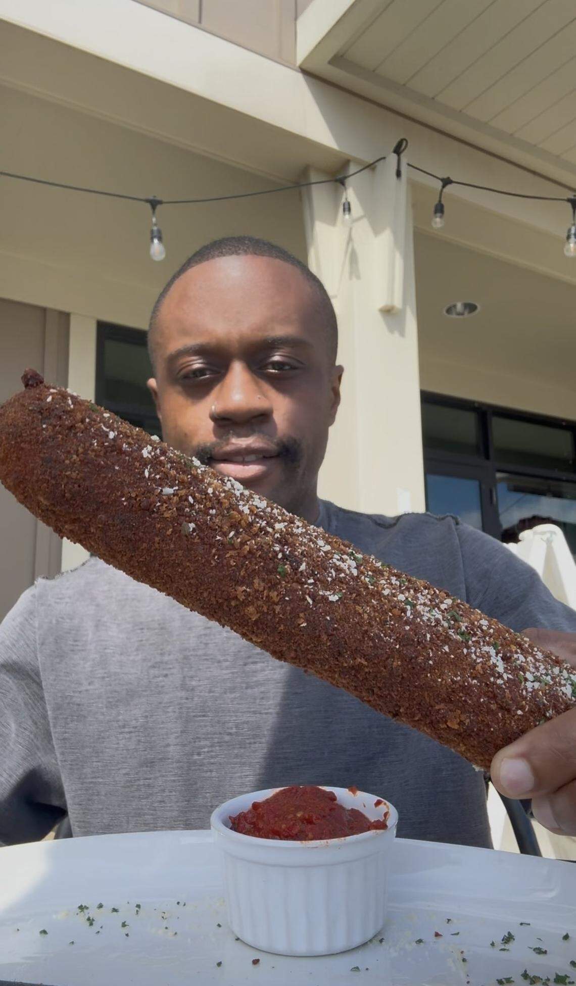 A man holding an exceptionally large, foot-long mozzarella stick coated in dark, crispy breading and herbs, held over a small bowl of marinara sauce.