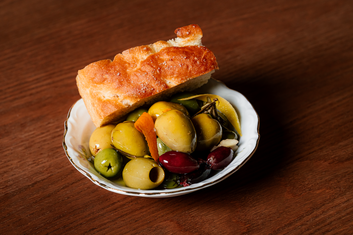 A close-up, slightly high-angle shot of a small, ornate white appetizer plate sitting on a dark wooden table. The plate contains a cluster of green and dark purple olives, glistening with oil; slices of orange zest and fresh rosemary sprigs tucked among the olives; and a thick, golden-brown slice of salted focaccia bread resting on top of the olives.