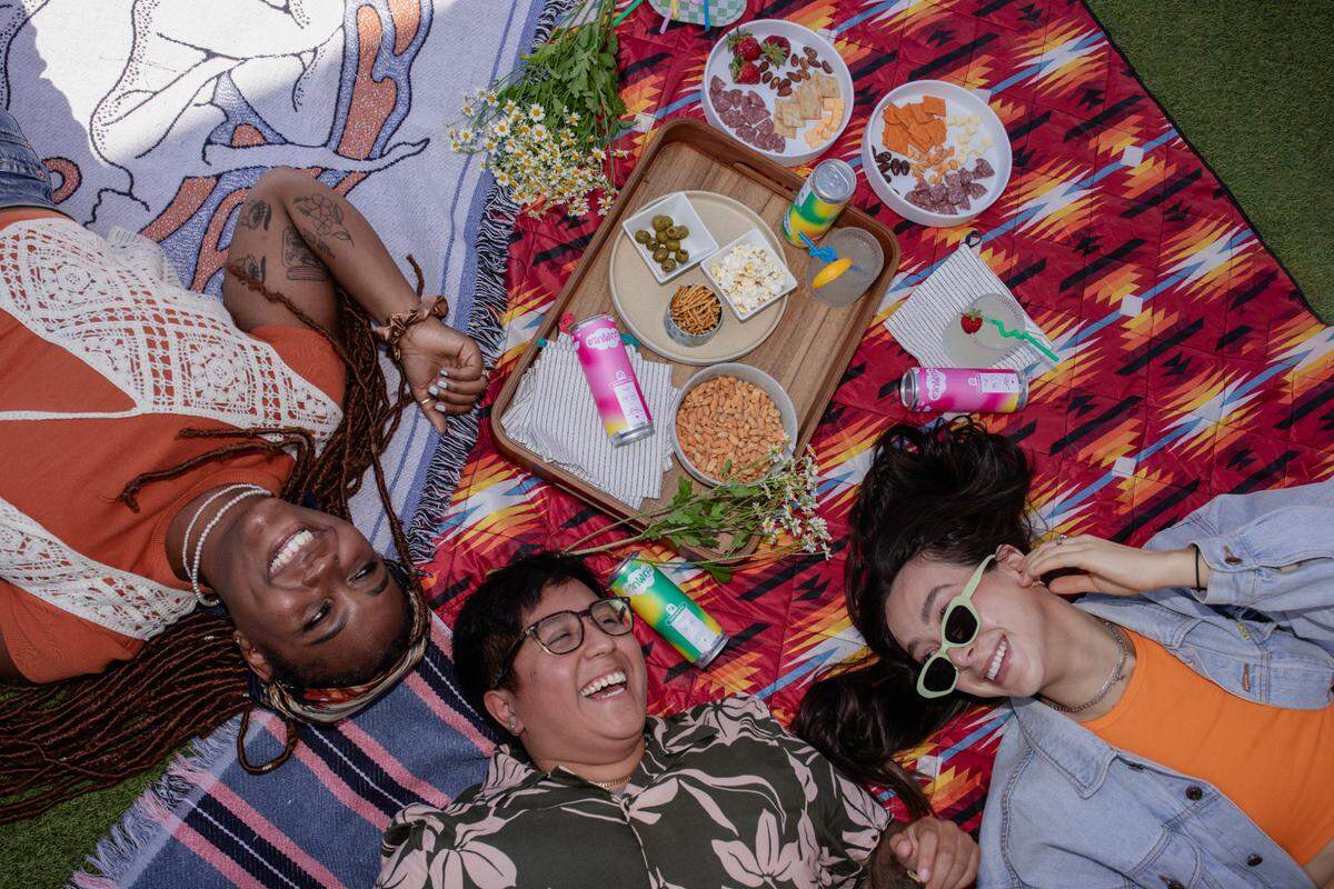 A joyful, overhead, high-angle shot of three people having a picnic, lying on their backs on vibrant, patterned blankets. Their heads are near the center, surrounding a wooden tray of food. The person on the left is smiling broadly, the person in the middle is laughing, and the person on the right smiles while wearing light green sunglasses. The tray holds bowls of charcuterie, nuts, olives, and pretzels, and is surrounded by colorful cans of beverages and a small bouquet of daisies.
