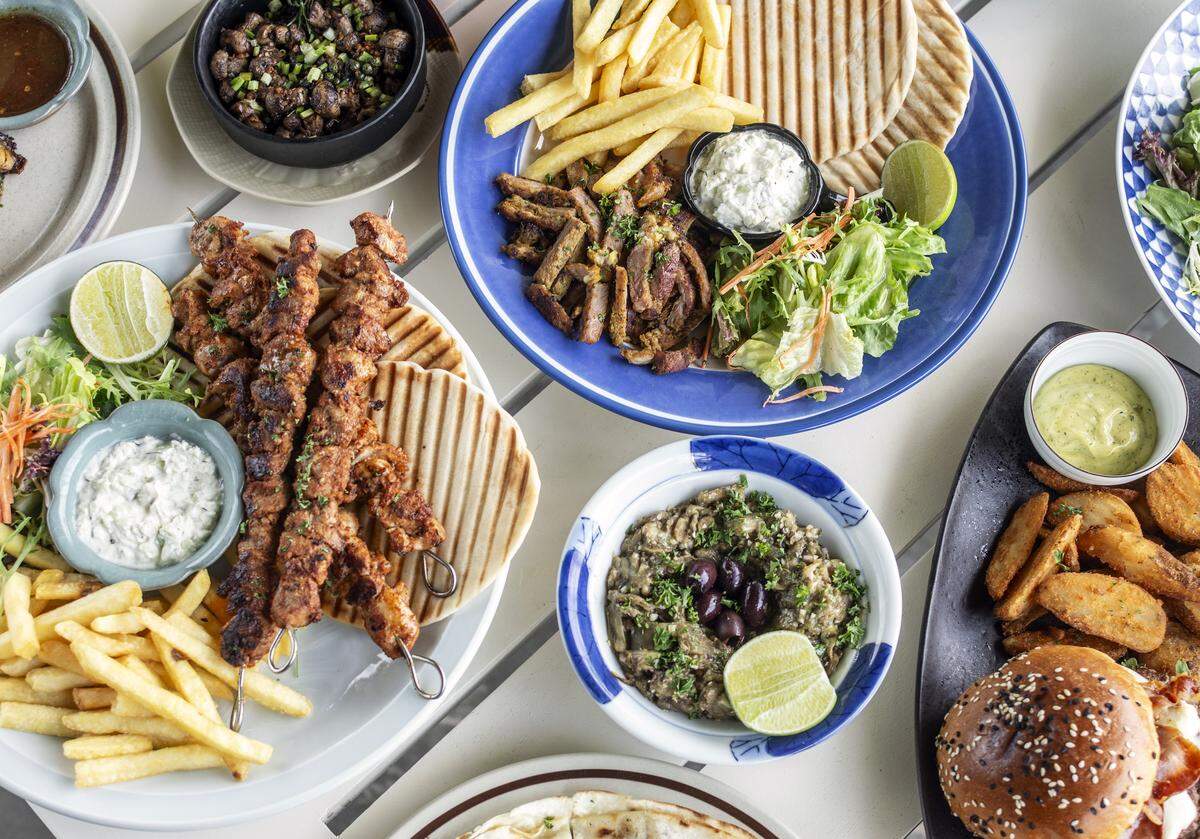 A vibrant, high-angle overhead shot of a table spread filled with a variety of Mediterranean and Greek dishes. The colorful presentation features several main plates and side dishes, including seasoned chicken souvlaki skewers, shaved pork gyros, grilled pita bread, golden french fries, and fresh side salads with lime wedges. Accompanying the platters are small bowls containing tzatziki, a roasted eggplant dip topped with kalamata olives, and a side of potato wedges with a burger.