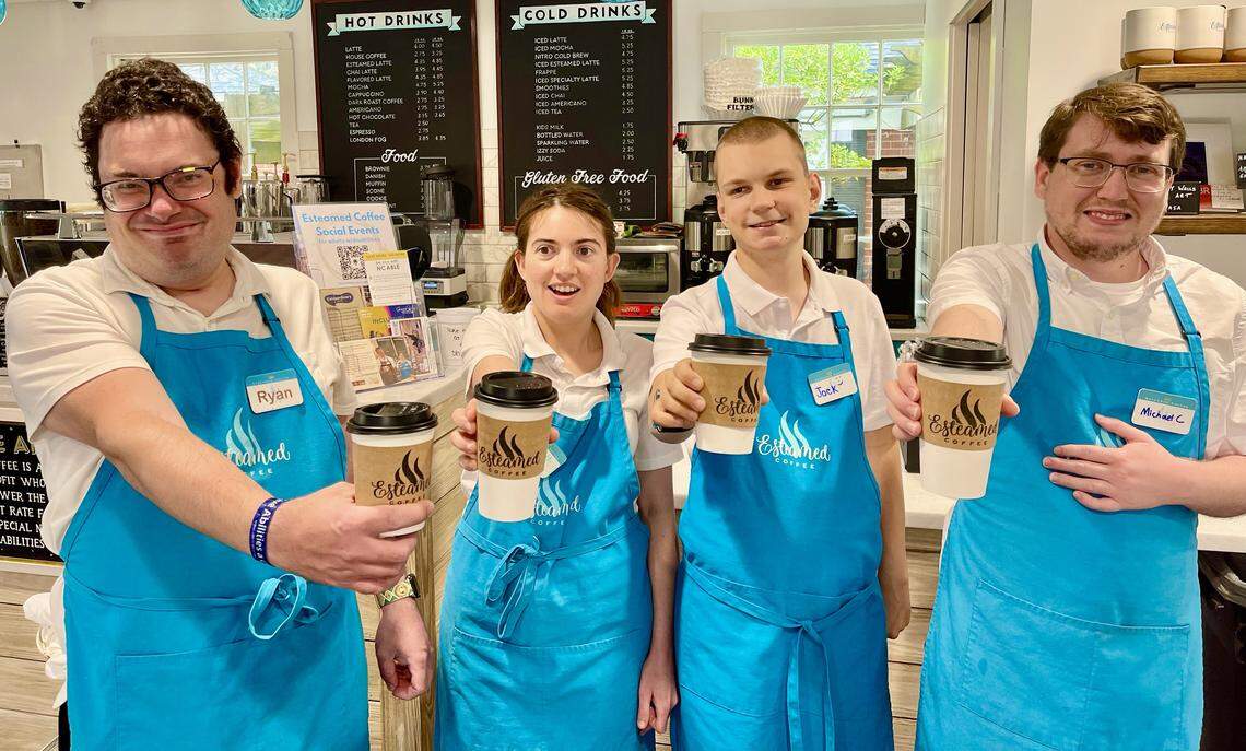 Four smiling baristas stand side-by-side behind a coffee counter, each holding out a branded coffee cup in a friendly toast toward the camera. They are all wearing white polo shirts and turquoise aprons, with the café’s chalkboard drink menu visible on the wall behind them.