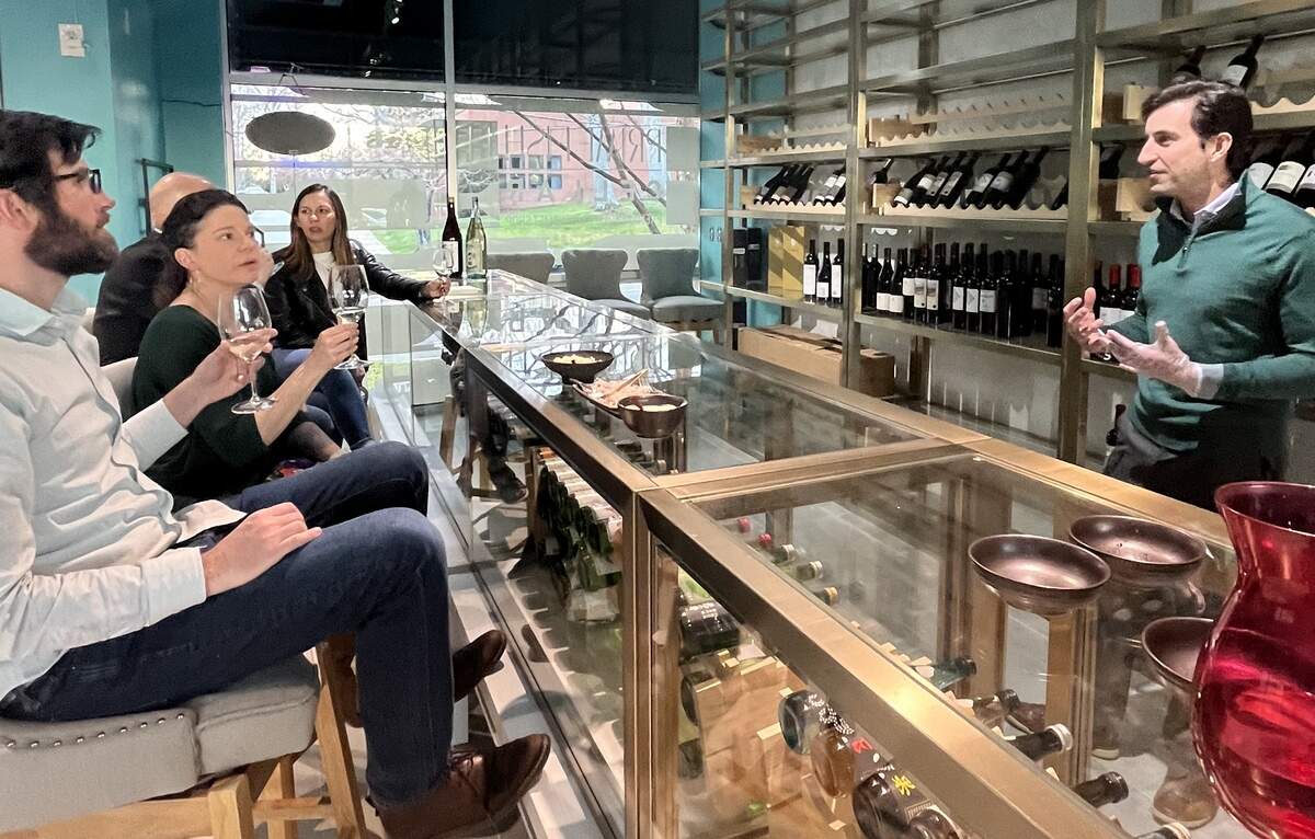 A group of people at a wine tasting in a modern bar. A man in a green sweater stands behind the counter, gesturing as he speaks to four seated patrons who are holding wine glasses and listening attentively. The bar has a glass display case, and shelves filled with wine bottles line the wall in the background.