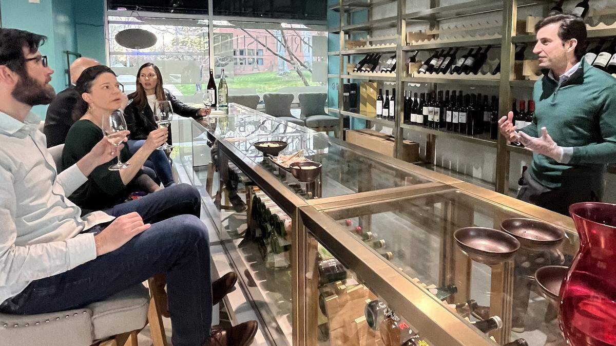 A group of people at a wine tasting in a modern bar. A man in a green sweater stands behind the counter, gesturing as he speaks to four seated patrons who are holding wine glasses and listening attentively. The bar has a glass display case, and shelves filled with wine bottles line the wall in the background.