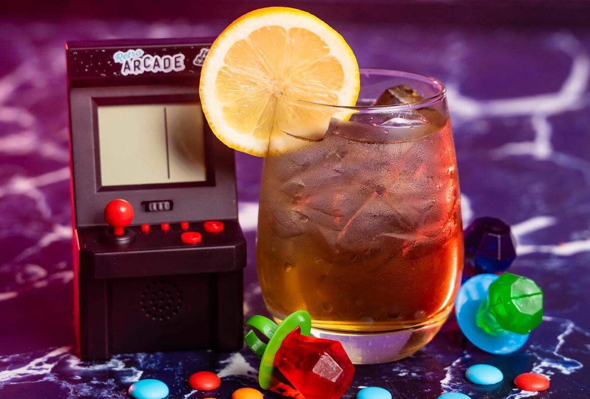 An iced cocktail garnished with a lemon wheel sits next to a miniature arcade game. Candy Ring Pops and other colorful candies are scattered on the table.