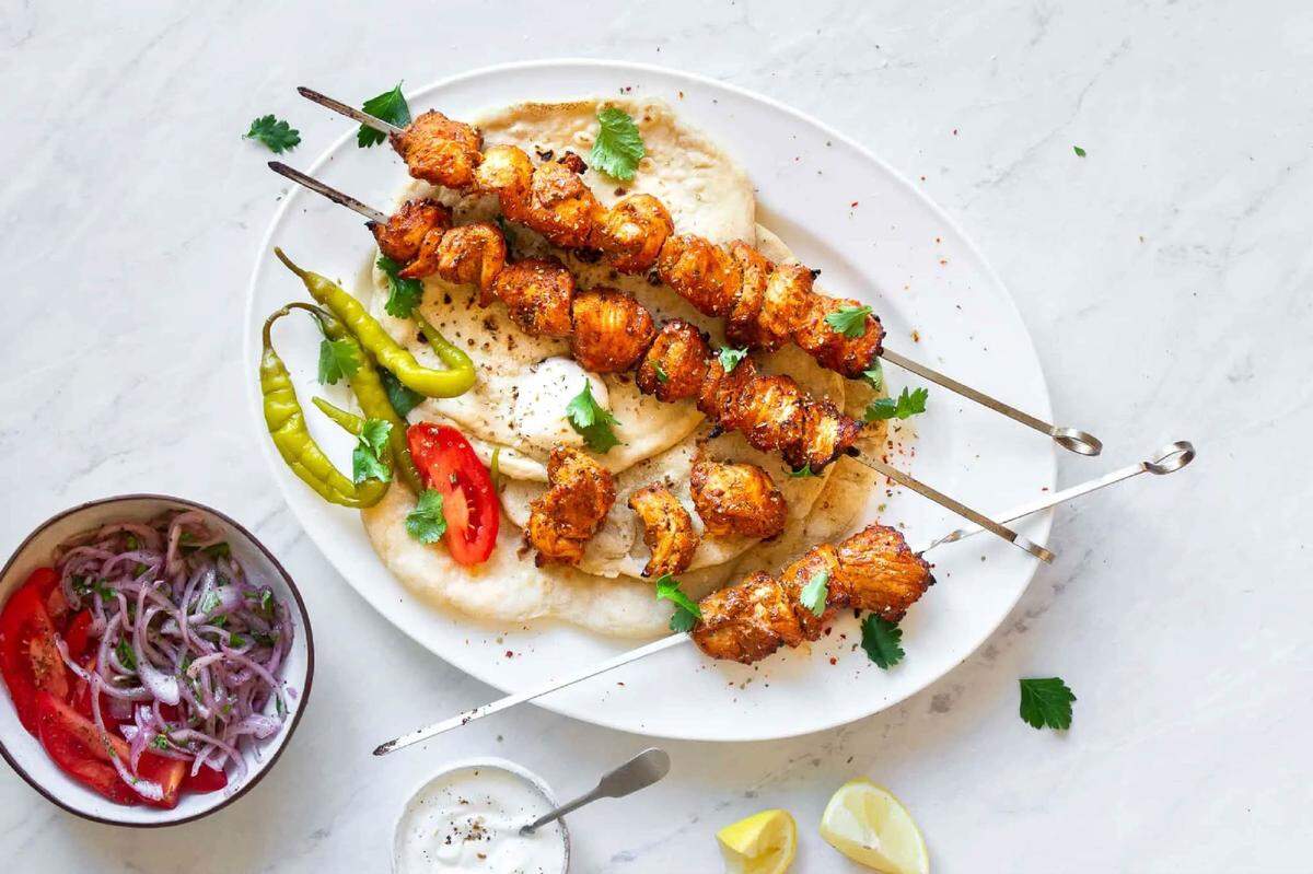 A top-down view of three metal skewers loaded with charred, seasoned chicken kababs resting on a bed of fluffy pita bread on a white platter. The dish is garnished with fresh cilantro leaves, sliced red tomatoes, and long green chili peppers. On the side, there is a small bowl of red onion and tomato salad, a small dish of white garlic sauce (toum), and lemon wedges, all arranged on a white marble surface.