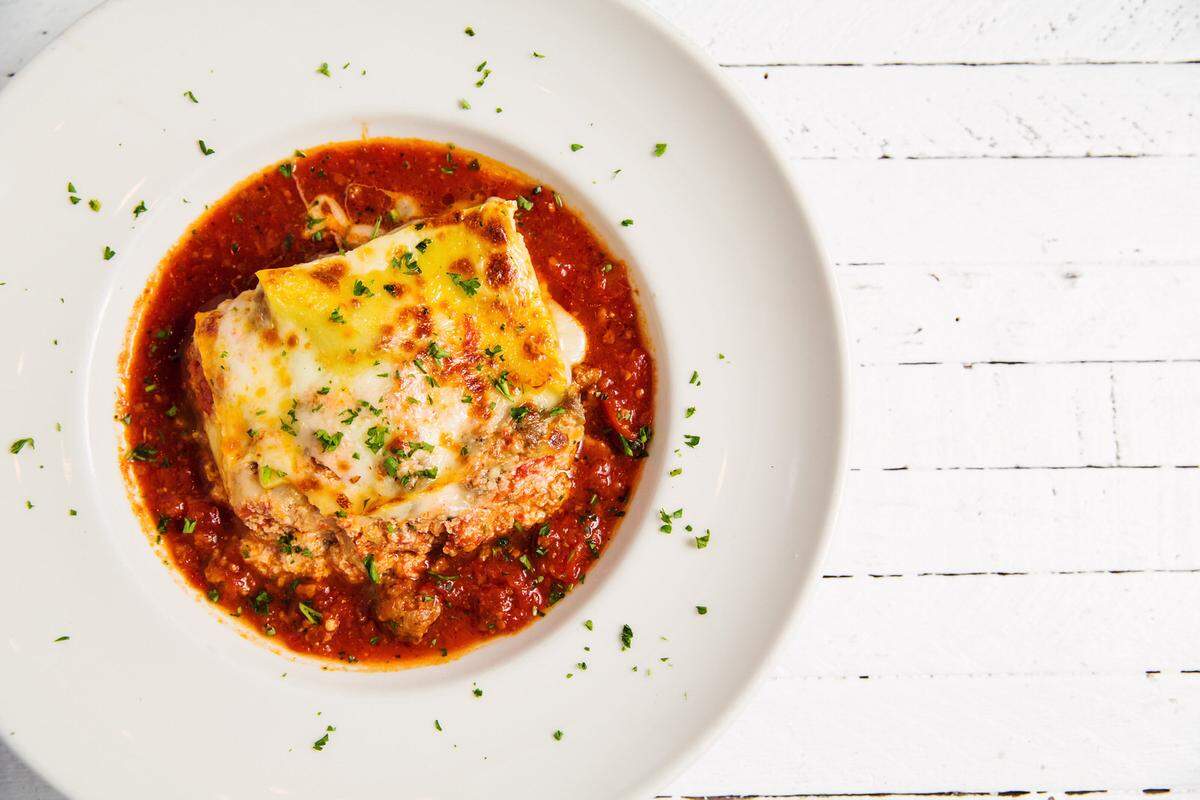 An overhead view of a portion of lasagna in a white bowl, covered in red meat sauce and melted cheese, and garnished with parsley.
