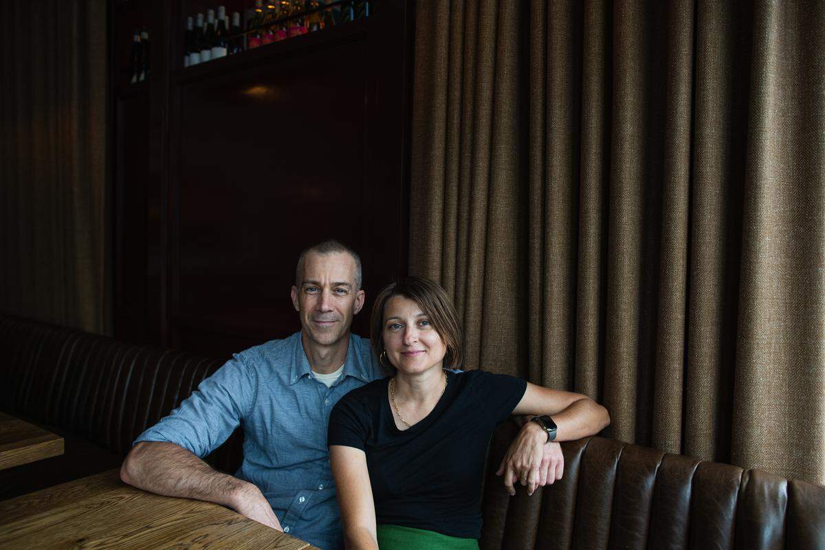 A portrait of Dean Neff and Lydia Clopton sitting together in a restaurant booth. Dean is wearing a blue button-down shirt, and Lydia is wearing a black t-shirt and a green skirt. They are positioned in front of a dark wood-paneled wall and heavy brown curtains, with several bottles of wine visible on a shelf in the background.