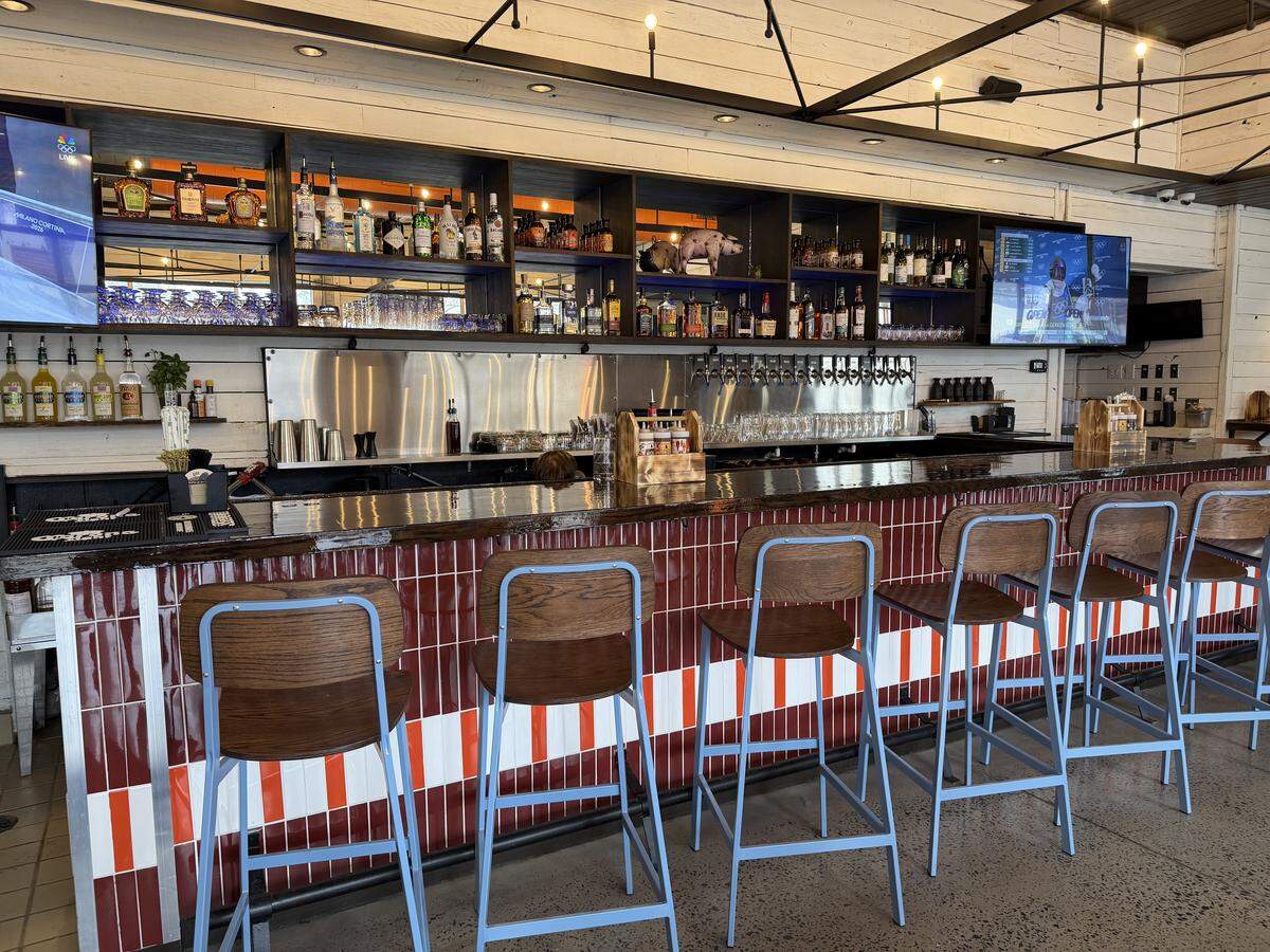 A long, polished dark wood bar with a base featuring vertical red and white striped tiles. Blue metal barstools with dark wood seats are lined up along the counter. Behind the bar, there is a stainless steel backsplash, multiple beer taps, and dark wood shelving stocked with various liquor bottles. Two televisions are mounted on the white wooden wall at either end of the shelving unit.