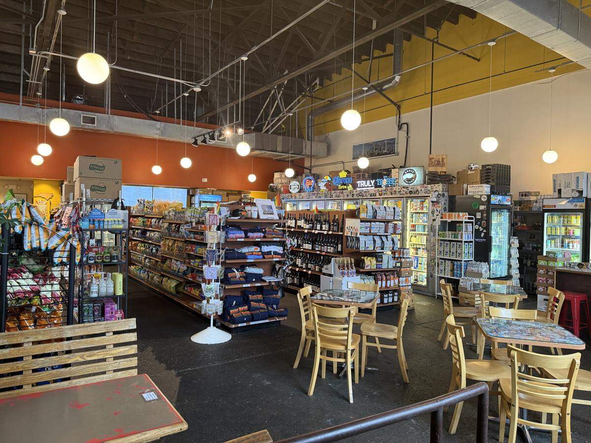 There are several small tables with wooden chairs in the foreground and to the right, suggesting it’s a place where customers can sit down and have a drink or a bite to eat. The walls are painted in bright orange and yellow, and the ceiling is high with exposed pipes and ductwork, giving it a somewhat industrial but casual feel.