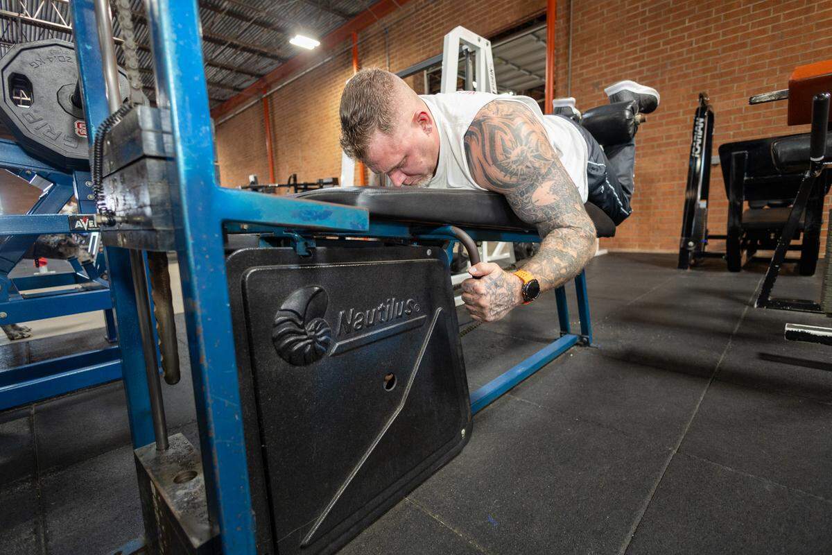 Prone gym user with a visible orange smartwatch performing a leg curl on a blue Nautilus machine. The black casing displays the Nautilus logo.