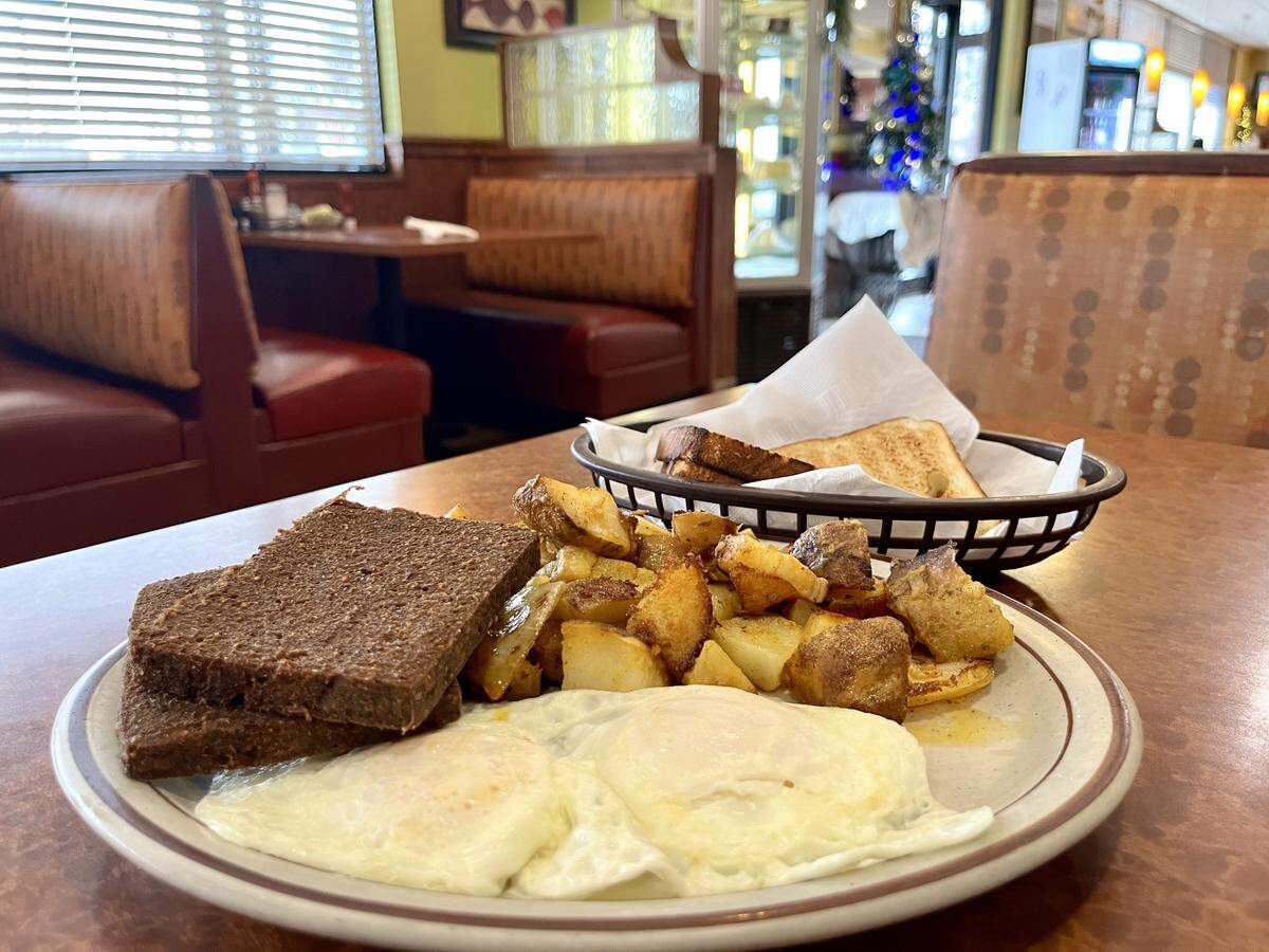 A plate of breakfast food featuring two sunny-side-up eggs, a generous serving of crispy home fries, and two slices of cooked livermush, set on a brown diner table. A basket of assorted toast is in the background. The diner interior shows red and brown vinyl booths and a glimpse of a holiday decoration.