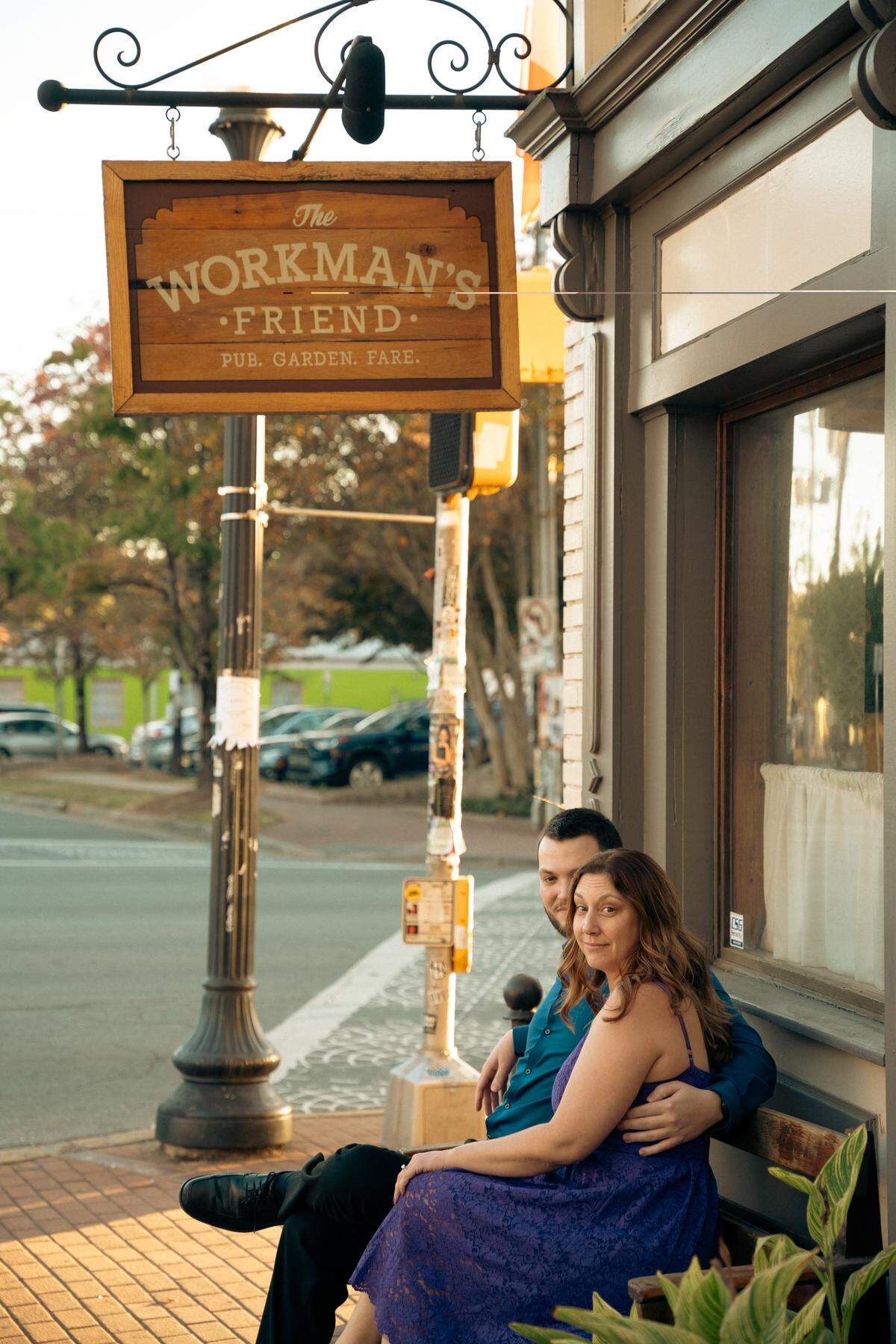 A man and woman sit close together on an outdoor wooden bench, smiling, under the hanging sign for “The Workman’s Friend” pub.