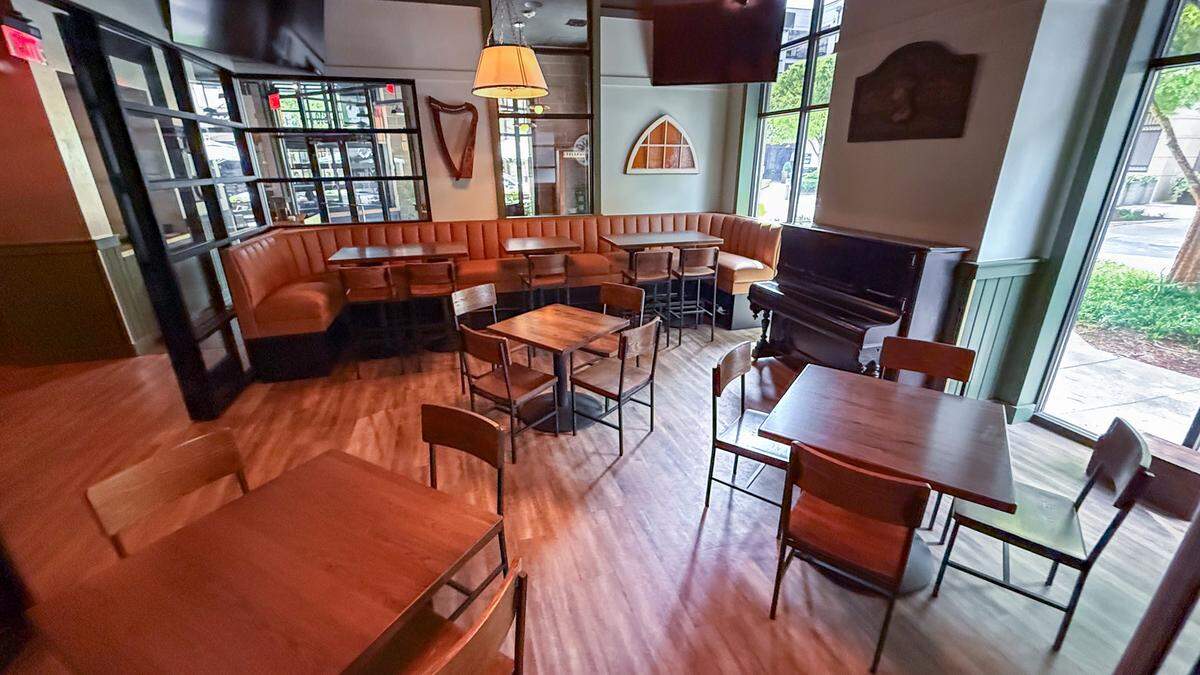 A cozy corner of the dining room featuring a long, L-shaped tan leather booth. Several small wooden tables with matching chairs are arranged in the space. Unique decor includes an Irish harp mounted on the wall and an upright dark wood piano tucked into the corner near a large window, adding a classic pub atmosphere.