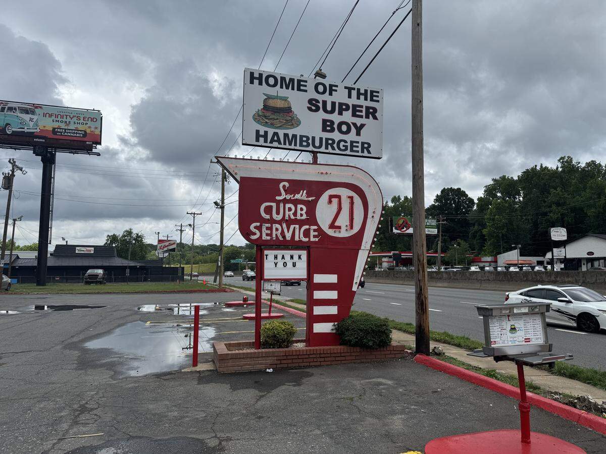 A vintage roadside sign for “South 21 Curb Service” next to a multi-lane highway under a cloudy sky. The main sign is shaped like a red arrow pointing downward, featuring a white circle with the number “21”. Above it, a rectangular white sign reads “HOME OF THE SUPER BOY HAMBURGER” with an illustration of a double-decker burger. A menu stand on a red pole sits on a patch of asphalt in the foreground.